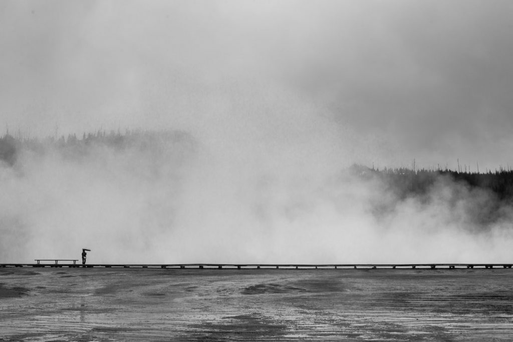 Person standing on boardwalk at Yellowstone National Park in the distance with fog rising in black and white